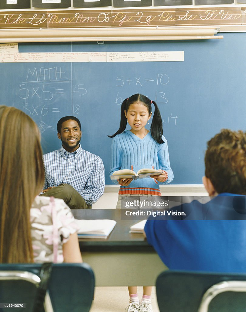 Young Girl Standing at the Front of a Classroom Reading From a Textbook