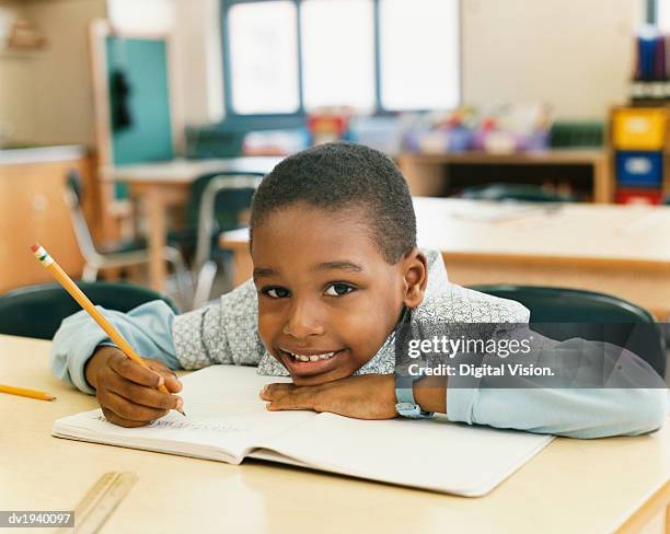 portrait of a smiling schoolboy sitting at a table and resting his head on a exercise book - workbook stock pictures, royalty-free photos & images