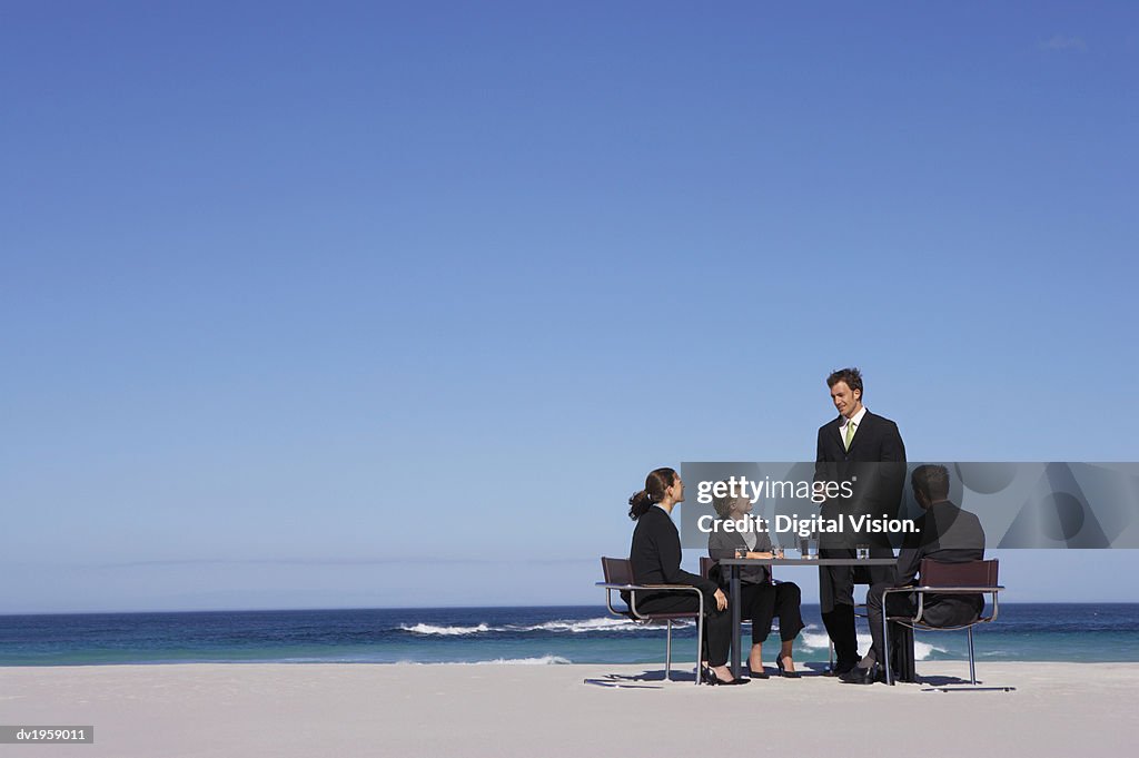 Four Well-Dressed Businessmen and Businesswomen Sit at a Table on a Beach, Having a Meeting