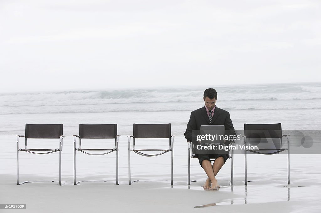 Businessman Sitting in a Line of Chairs on a Beach Working on His Laptop