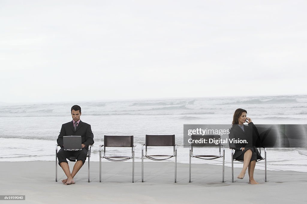 Businesswoman and Businessman Sitting Apart on a Beach Using a Laptop and a Mobile Phone