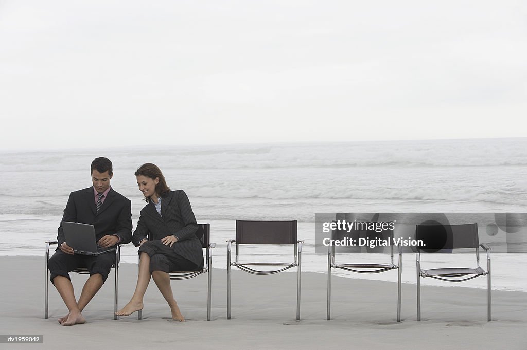 Businessman and Businesswoman Sit at the End of a Row of Chairs on a Beach, Using a Laptop