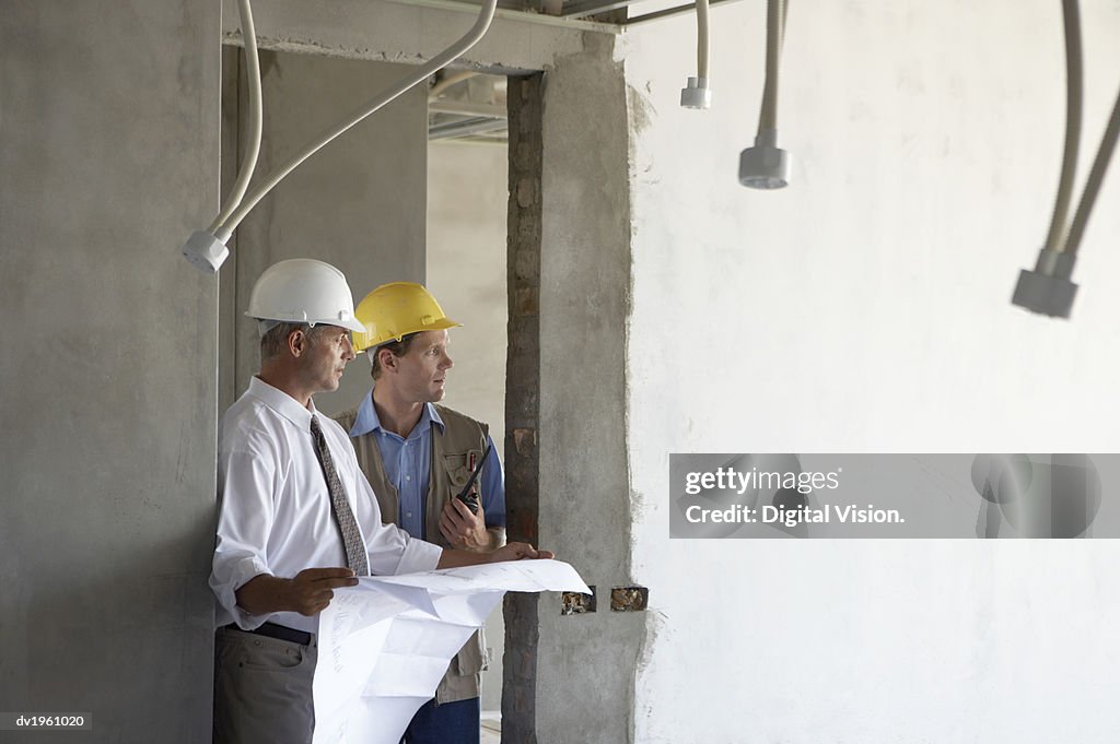 Two Male Construction Workers Stand in an Incomplete Building Discussing a Blueprint