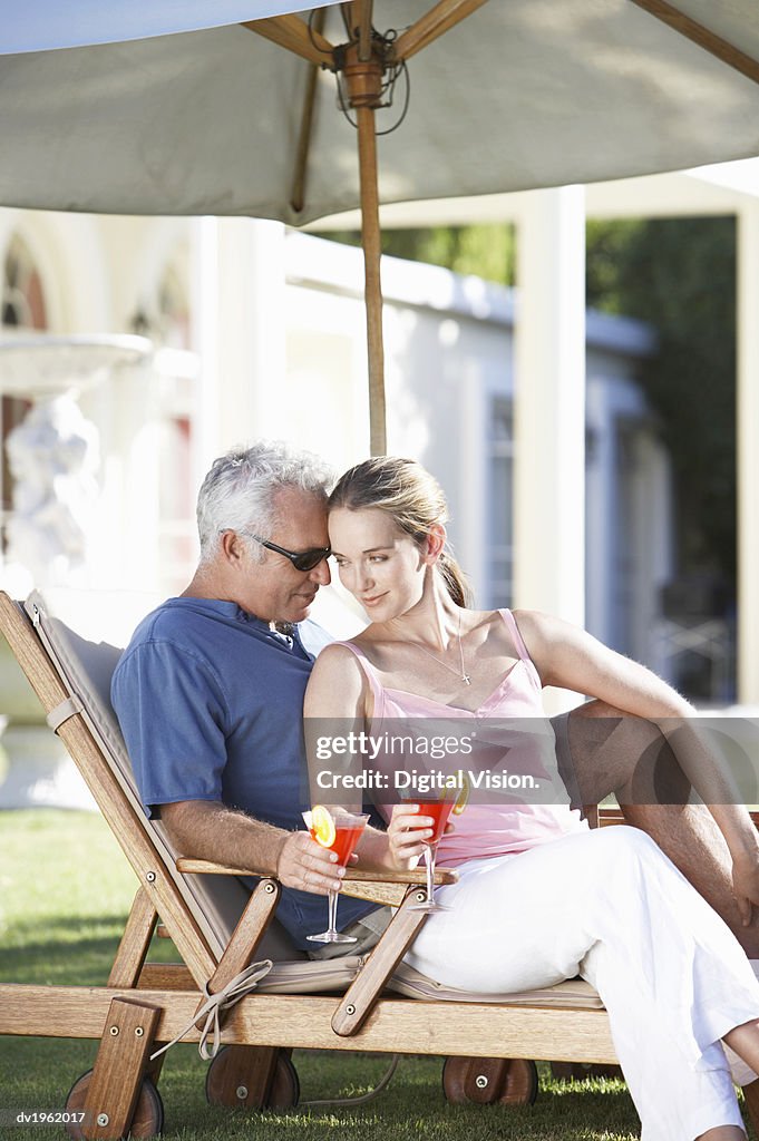 Affectionate Couple Sit Under a Parasol on a Sun Lounger Holding Cocktail Glasses