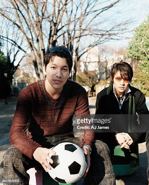 two young men sitting in a park, with one man holding a football - v hals stockfoto's en -beelden