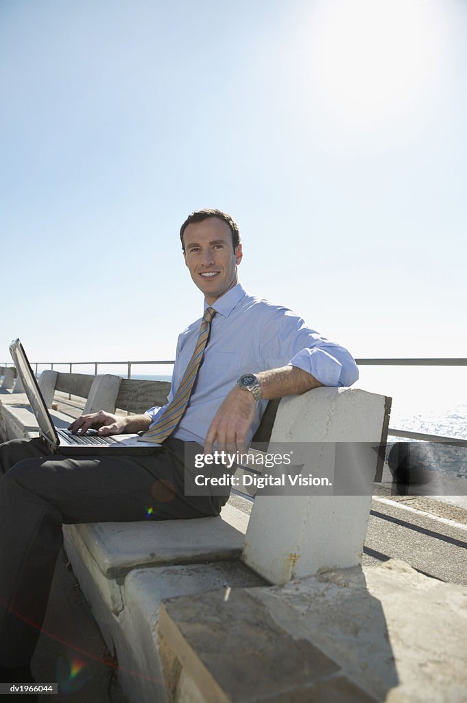 Smiling Businessman Sitting on a Waterfront With a Laptop