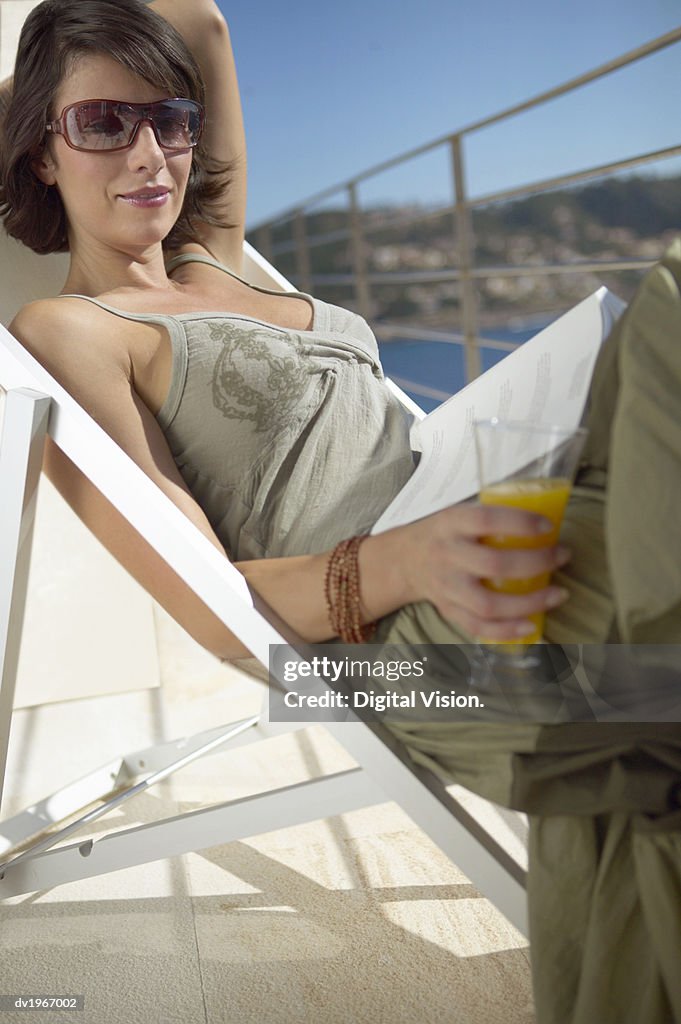 Woman Lies on a Sun Lounger on Her Balcony, Holding a Glass of Orange Juice