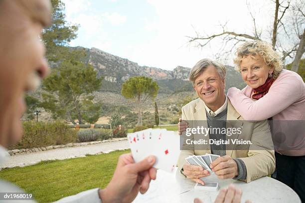 two senior men sit at a table in the countryside playing a card game, their friend watching - card table stock pictures, royalty-free photos & images