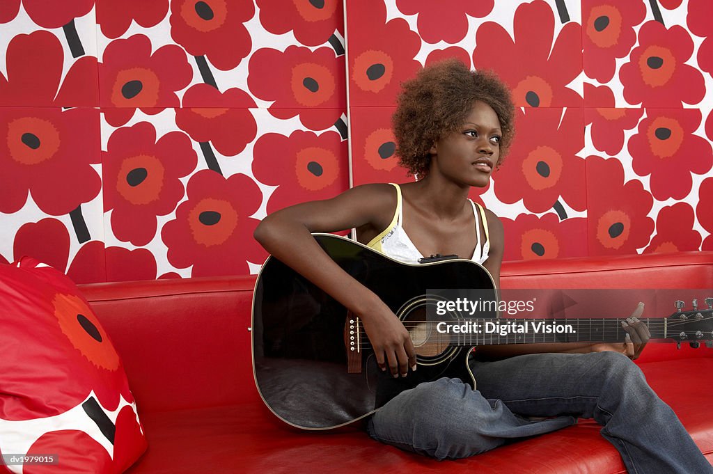 Young Woman Playing an Acoustic Guitar on a Red Leather Sofa Against Floral Wallpaper