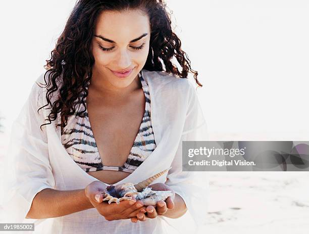 young woman sits on the beach looking down at seashells she is holding - down blouse stockfoto's en -beelden