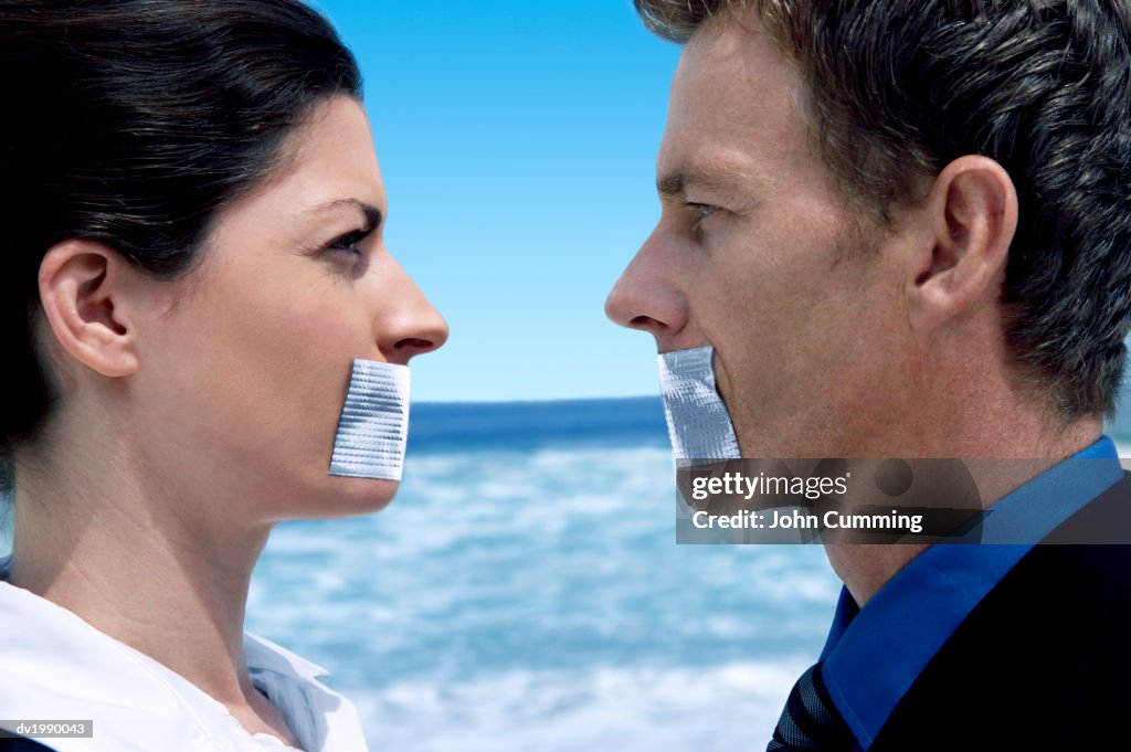 Businessman and Businesswoman Standing Face to Face on a Beach With Their Mouths Covered in Duct Tape