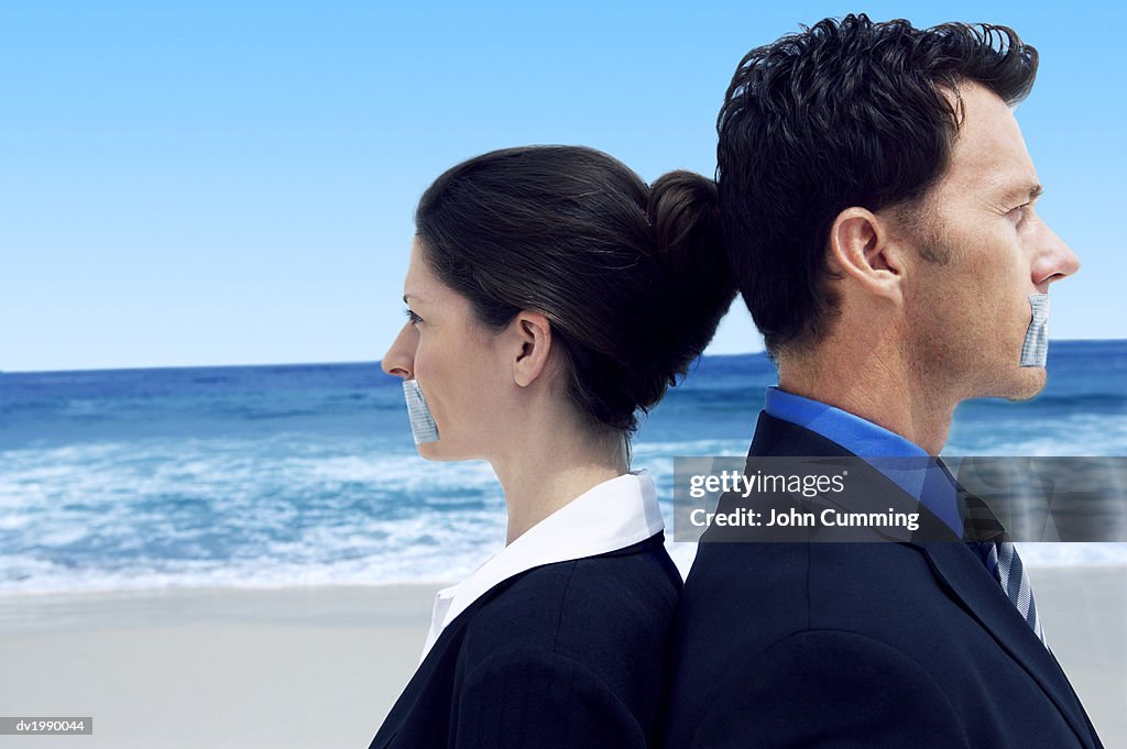Two Business Executives Standing Back to Back on a Beach With Their Mouths Covered by Duct Tape