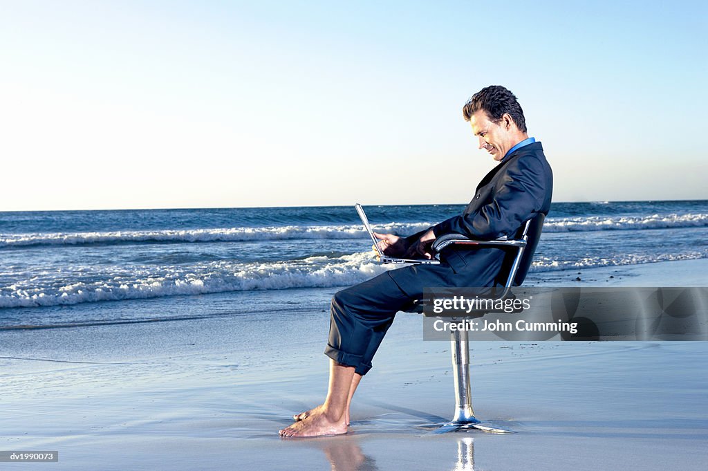 Businessman Sitting on a Beach by the Sea in a Swivel Chair and Working on His Laptop