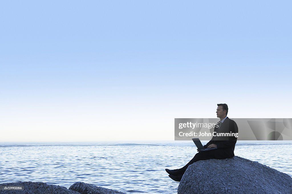Tranquil Businessman Sitting on a Rock by the Sea and Using a Laptop Computer