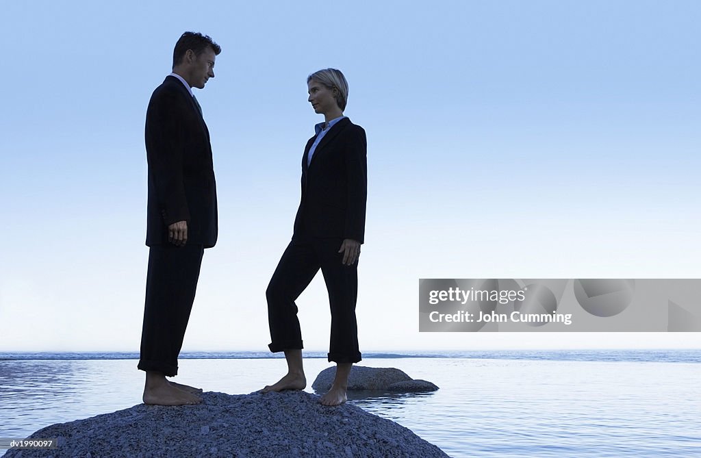 Two Barefoot Business Executives Standing Face to Face on a Rock in the Sea