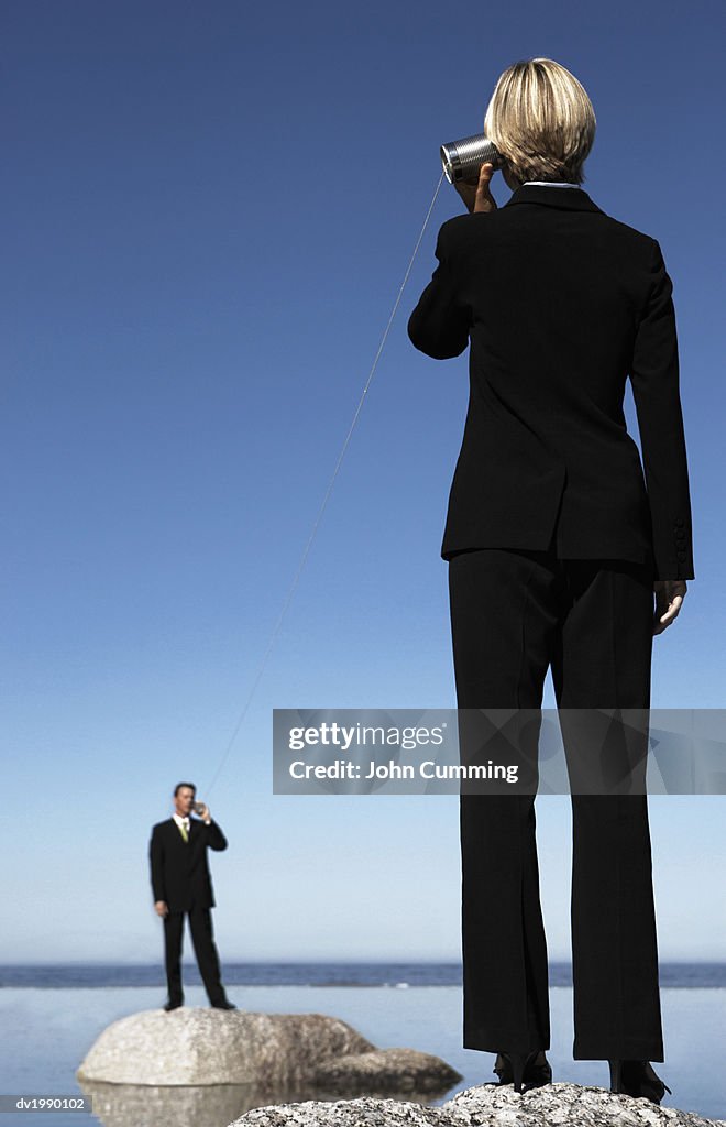 Business Executives Standing on Rocks in the Sea, Communicating Through Two Tin Cans Connected by String