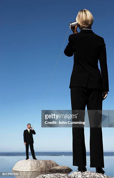 business executives standing on rocks in the sea, communicating through two tin cans connected by string - string - fotografias e filmes do acervo