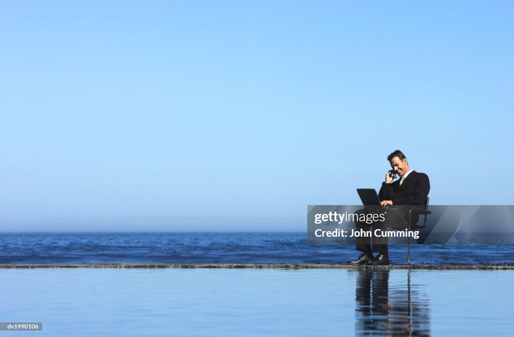 Businessman Talking on a Mobile Phone and Using a Laptop Computer by the Sea