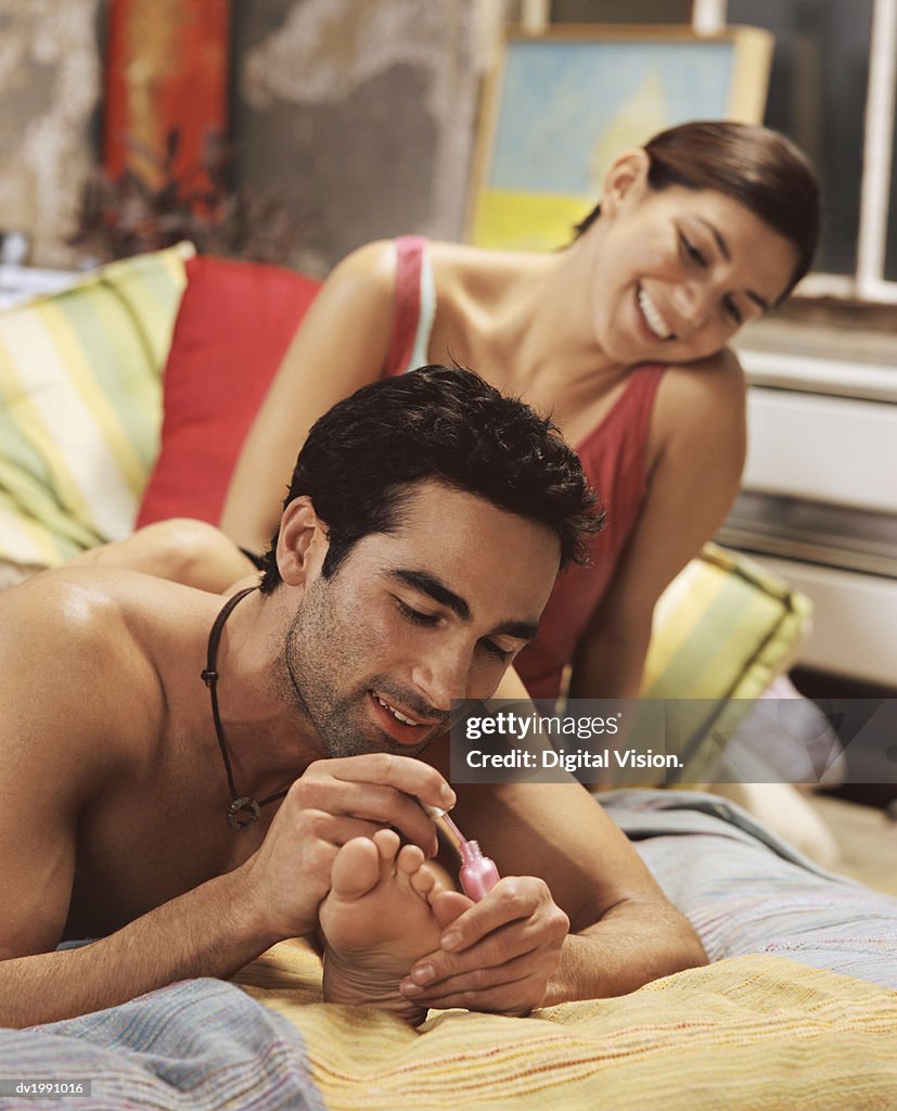Young Man Lies on a Bed, Painting His Girlfriend's Toe