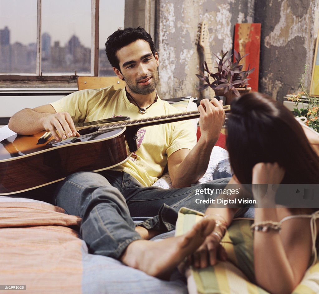 Twentysomething Couple on a Bed in an Apartment, Man Playing an Acoustic Guitar