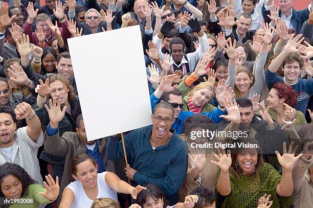 large crowd of people with their hands raised in the air with one man holding a blank placard - group of people protesting with placards foto e immagini stock