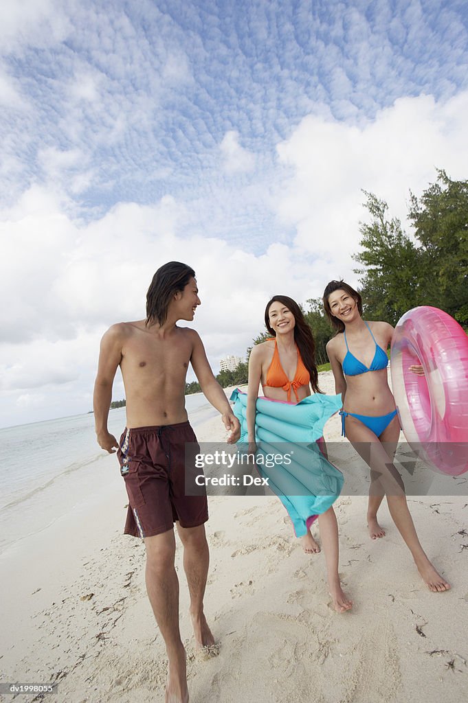 Three Young Adults Walking Along a Beach and Carrying Inflatables
