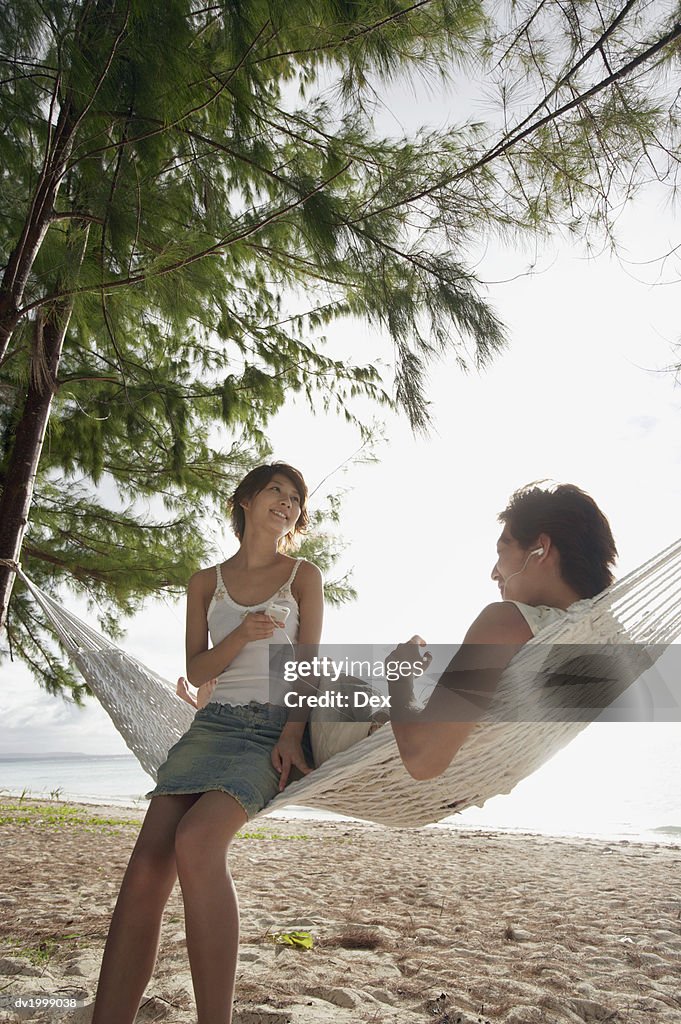 Young Couple Sit on a Hammock on the Beach Listening to Music on Headphones