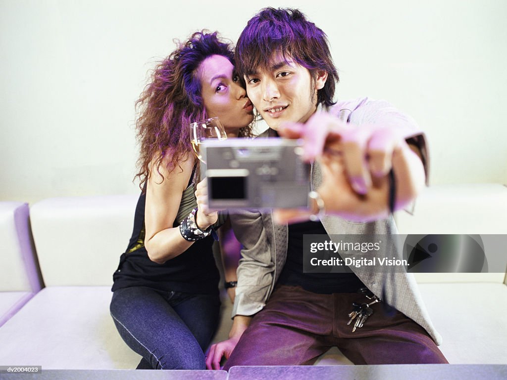Couple Sitting on a Sofa, Man Taking a Photograph of Themselves With a Digital camera Whilst The Woman is Kissing His Cheek