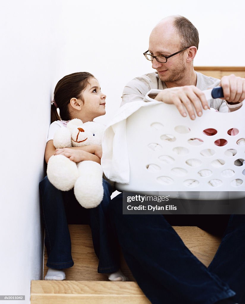 Man Sits on the Stairs With a Laundry Basket Next to His Young Daughter Holding a Teddy Bear