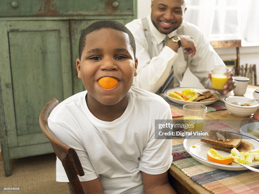 Boy With an Orange in His Mouth at Breakfast and His Father in the Background