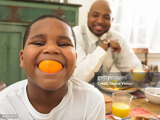 boy with an orange in his mouth at breakfast and his father in the background - child eating a fruit stock pictures, royalty-free photos & images