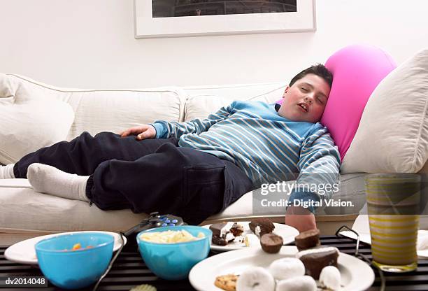 young, overweight boy sleeps on a sofa next to a table of crisps and biscuits - unhealthy living stock pictures, royalty-free photos & images