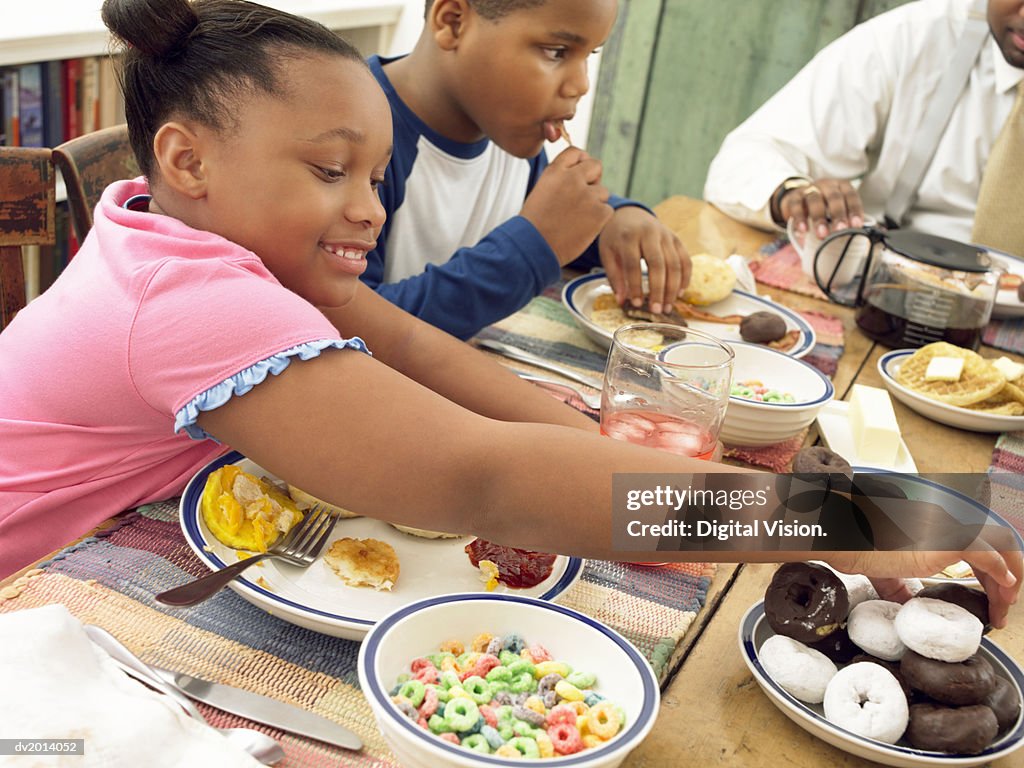 Young Girl Sits Next to Her Brother at the Breakfast Table With a Plate of Fried Food, Picking up a Doughnut