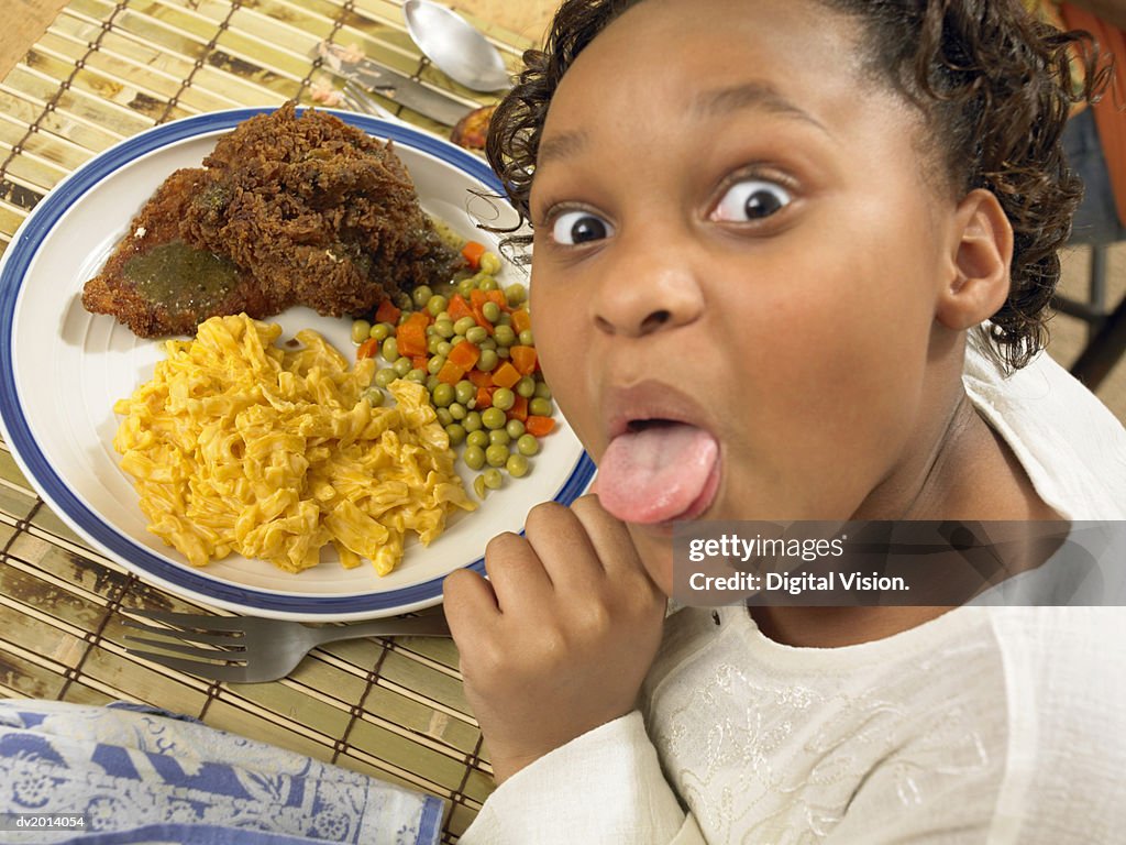 Young Girl Sits With a Plate of Fried Chicken and Macaroni Cheese, Sticking Her Tongue Out at the Camera