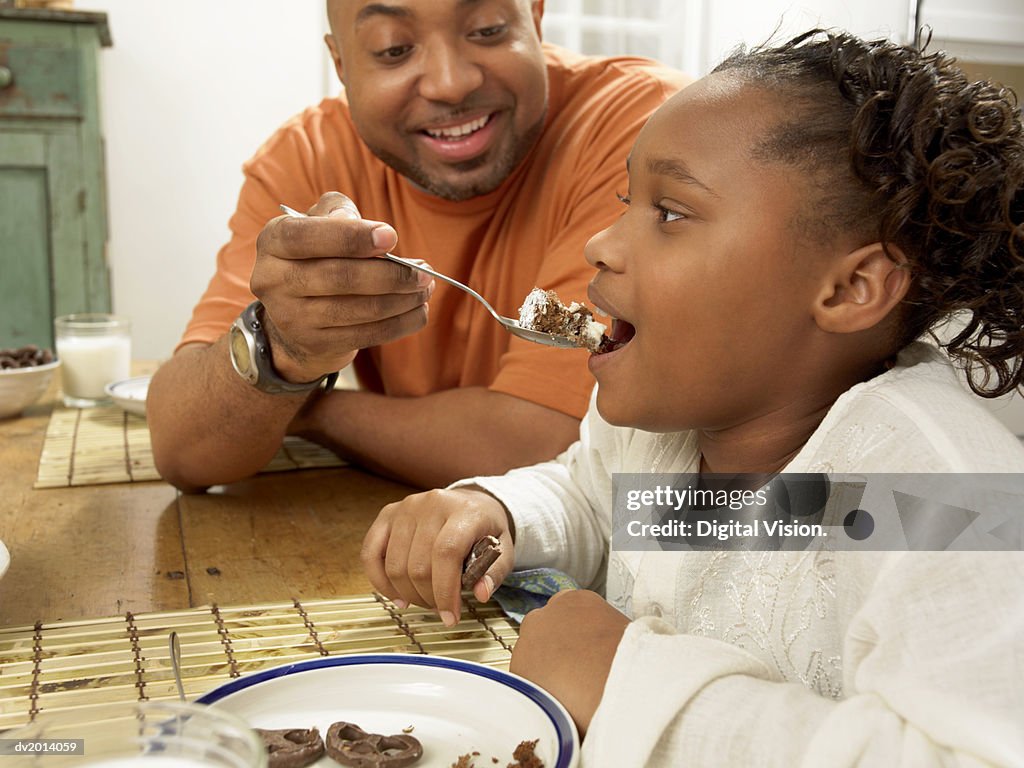 Man Sits at the Table With His Young Daughter, Feeding Her Cake From a Spoon