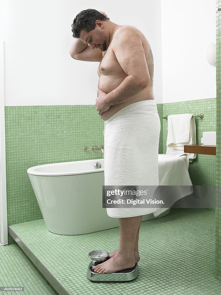 Overweight Man Wrapped in a Towel Stands on a Pair of Scales in a Bathroom