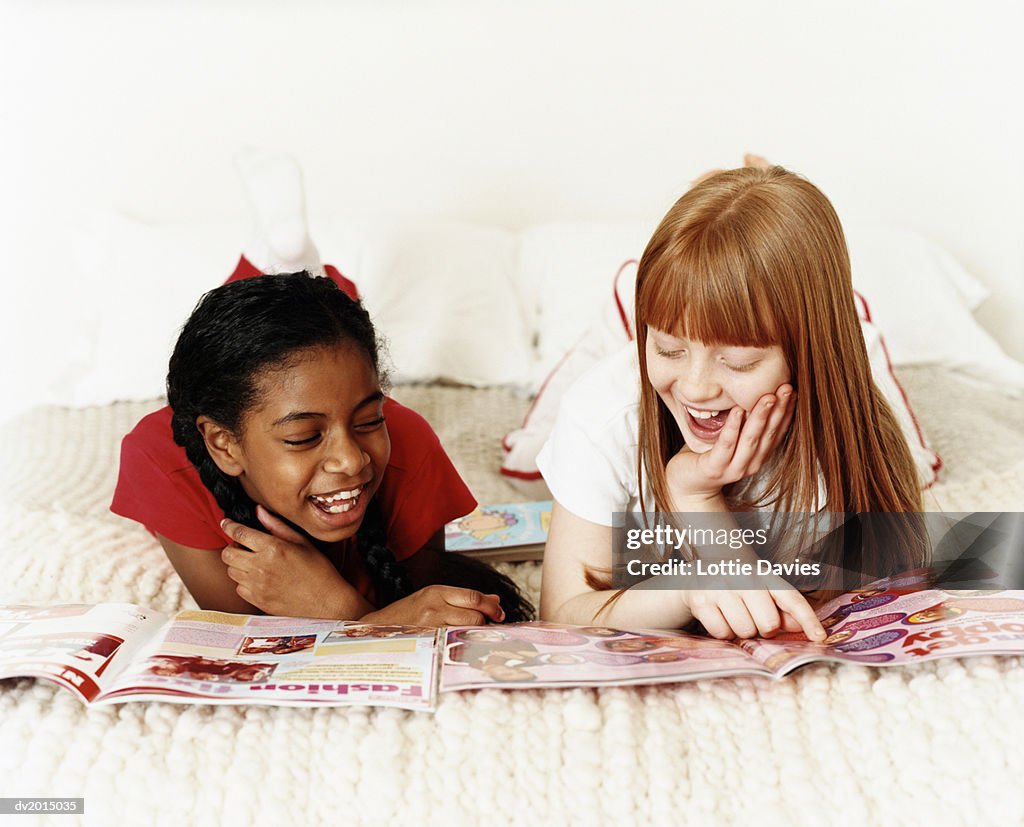 Two Teenage Girls Lying on Bed Reading Magazines, One Girl Pointing at an Image