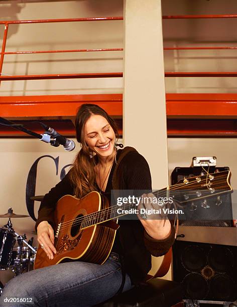 young woman sits by a microphone stand on a stage, playing a guitar and smiling - guitar stand stock pictures, royalty-free photos & images