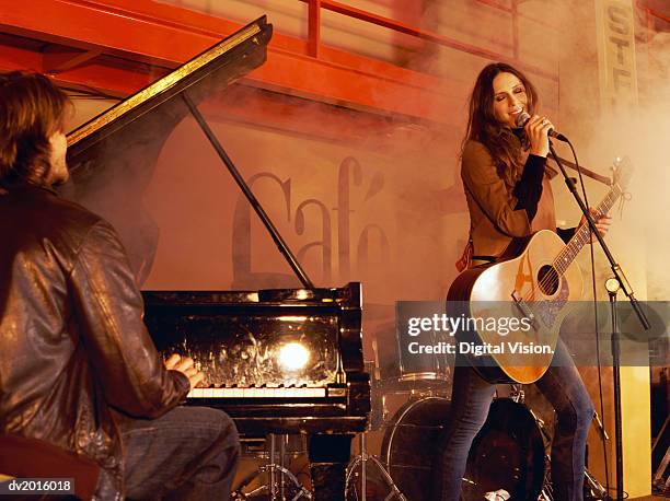 man playing piano and a woman holding an acoustic guitar and singing, on a smokey stage - bands stockfoto's en -beelden