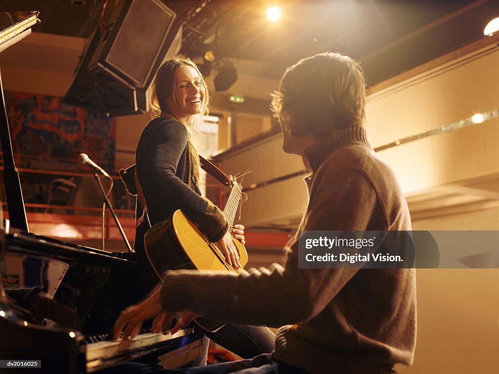 Woman Holding an Acoustic Guitar Leaning Against a Piano and Smiling at a Male Pianist