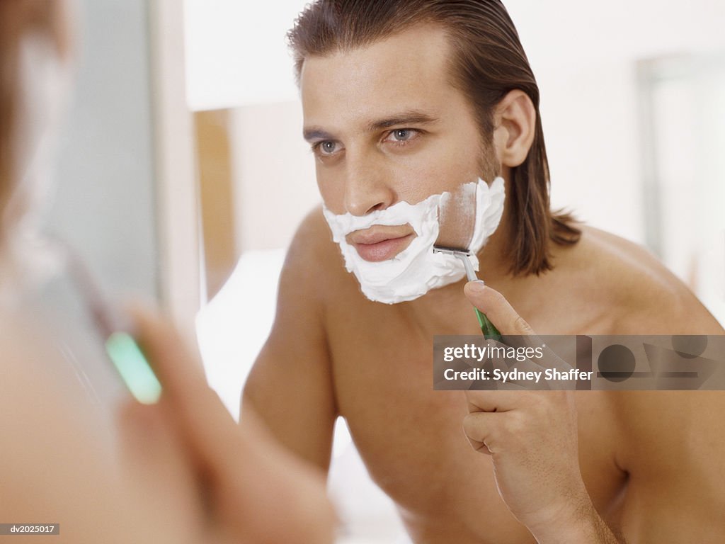 Man Looks in a Bathroom Mirror, Shaving His Chin With Shaving Foam and a Razor