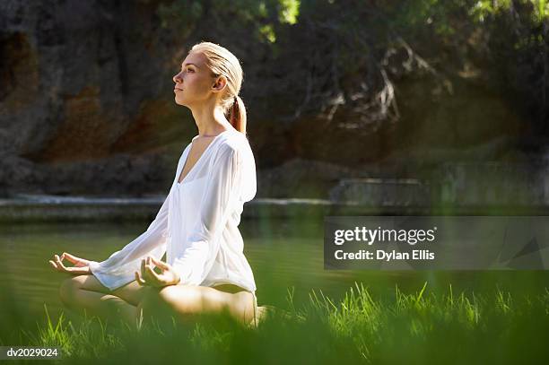 young woman sitting in the lotus position by a lake - ellis stock-fotos und bilder