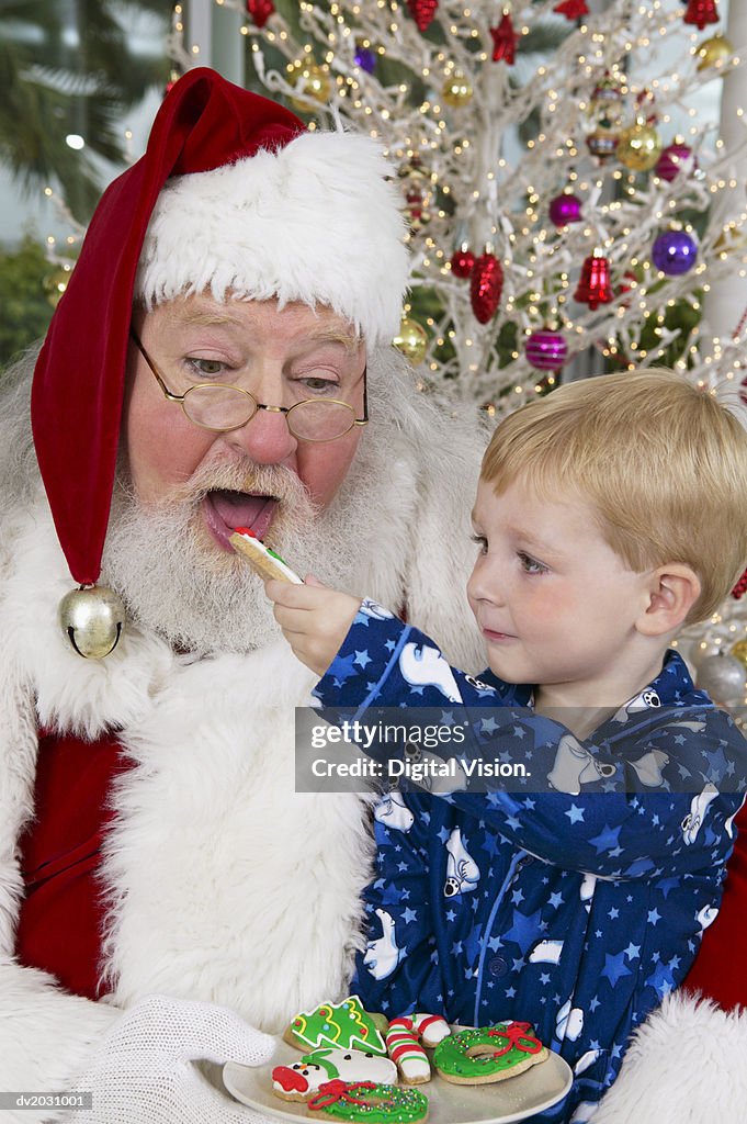 Young Boy Sits on Father Christmas' Lap, Feeding Him a Cookie