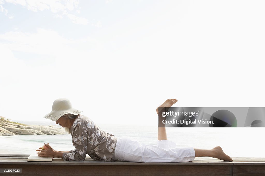 Senior Woman Lying on Wooden Decking on the Coast and Reading a Book