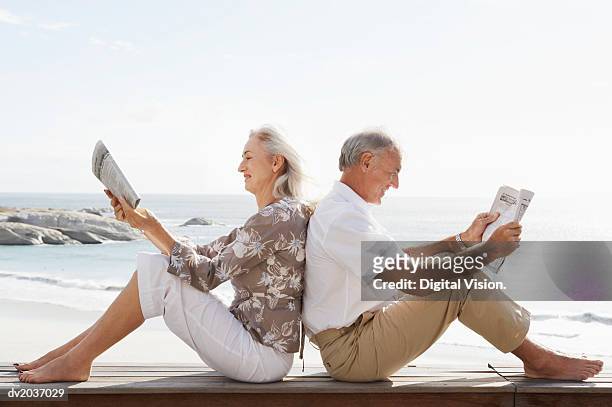 couple reading newspapers sitting back to back on wooden decking on the coast - beach deck stock pictures, royalty-free photos & images