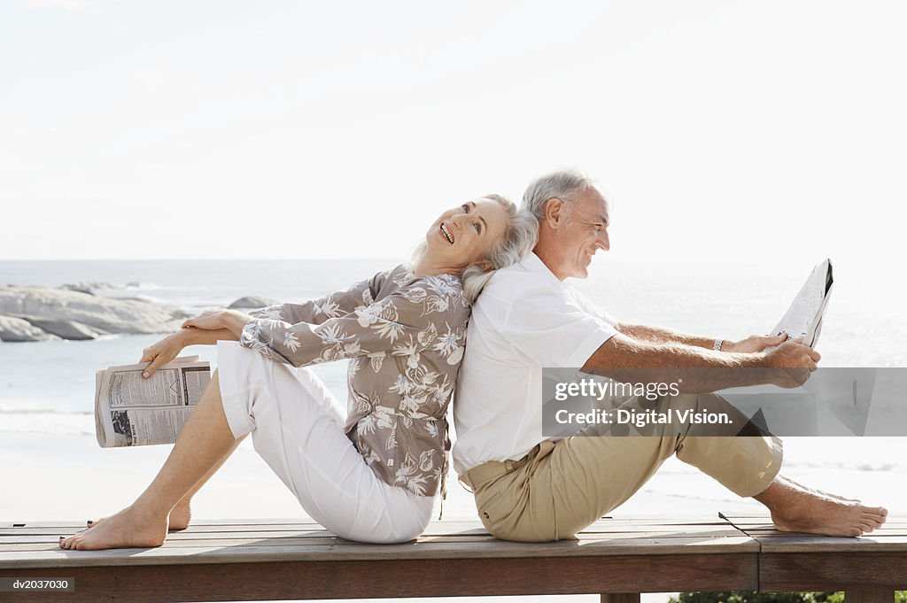 Couple With Newspapers Sitting Back to Back on Wooden Decking on the Coast