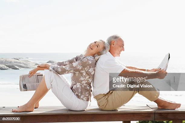 couple with newspapers sitting back to back on wooden decking on the coast - barefoot couples stock pictures, royalty-free photos & images
