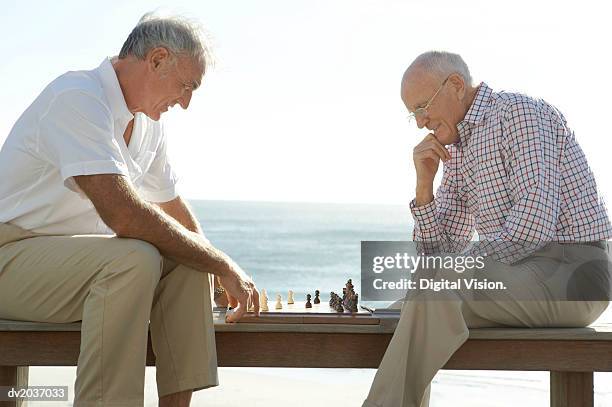 two senior men playing chess, with the sea in the background - kurzärmlig stock-fotos und bilder