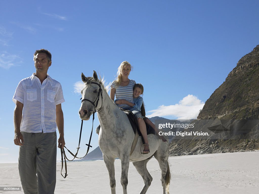 Mother and Daughter Riding a White Horse on the Beach With the Father Holding the Reins