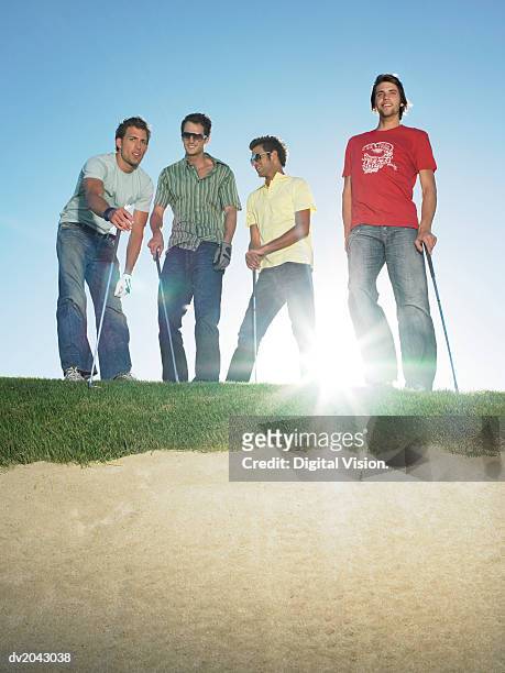 four young men standing at the edge of a sand bunker holding golf clubs - golf bunker low angle stock pictures, royalty-free photos & images